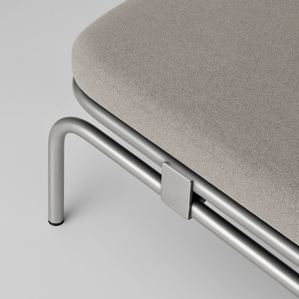 Détail d’un meuble design à Seraing avec structure en métal brossé et assise en tissu gris texturé, photographié en gros plan sur fond uni.